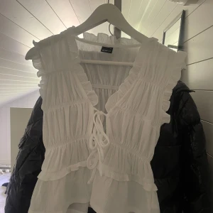 Vit volangblus från Gina Tricot XS - Helt oanvänd endast testad! Supersöt vit blus från Gina Tricot i storlek XS. Toppen har volangdetaljer, smockad struktur och knytning framtill. Ärmlös modell med v-ringning och lätt, luftig känsla – perfekt för varma dagar eller när du vill känna dig extra söt.