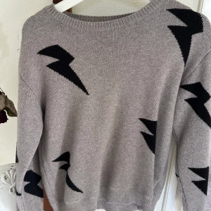 Grå stickad tröja med svarta blixtar - Grå stickad tröja med coola svarta blixtmönster över hela. Rund halsringning och långa ärmar. Perfekt för dig som vill sticka ut lite extra med en unik design och ändå hålla det stilrent.