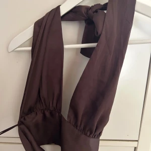 Brun omlottopp från Gina Tricot - Säljer en brun omlottopp från Gina Tricot i mjuk polyester. Toppen har breda axelband som knyts i nacken och en snygg omlottkänsla framtill. Perfekt för dig som gillar trendiga och stilrena plagg med en twist.