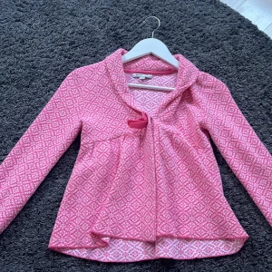 Rosa stickad kofta med rosett - Supersöt rosa och vit stickad kofta från My wear med mönster och en fin rosett framtill. Koftan har lång ärm, lätt utställd passform och mjukt material som känns skönt mot huden. Perfekt för dig som gillar färg och detaljer! Storlek 146/152