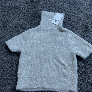 Glittrig polotröja från Zara - Supermjuk och glittrig polotröja från Zara i ljusgrått med korta ärmar och hög krage. Tröjan har en fluffig, stickad känsla och skimrar snyggt i ljuset.