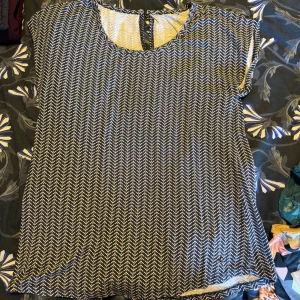 Svartvit mönstrad blus med rund hals - Säljer en svartvit blus med zigzag-mönster och rund hals. Toppen har korta ärmar och en liten knappdetalj i nacken. Materialet känns lätt och luftigt, perfekt för dig som gillar grafiska mönster och enkel stil.