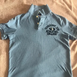 Blå pikétröja Superdry Athletics - Blå pikétröja från Superdry med broderad Athletics College-logga på bröstet och två knappar vid kragen. Klassisk kortärmad modell i mjuk bomull, perfekt för en sportig och chill stil. Snyggt uppvikt krage och stilren design.