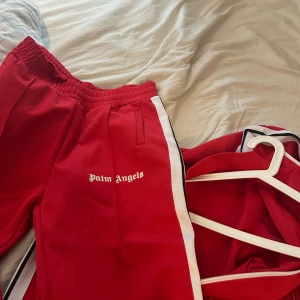 Palm Angels röd tracksuit med vita ränder - Palm Angels tracksuit i rött med vita och svarta ränder längs ärmar och ben. Setet består av zip-jacka och byxa med elastisk midja, båda med Palm Angels-logga tryckt på bröst och lår. Sportig look och bekväm passform, perfekt för streetwear. Storlek XL passar L 