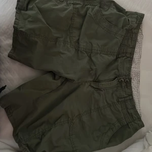 Gröna cargo shorts med fickor - Säljer ett par gröna cargo shorts med flera praktiska fickor både fram och bak. Shortsens midja har knapp och dragkedja, och materialet känns lätt och bekvämt. Perfekta för varma dagar och chill häng utomhus.