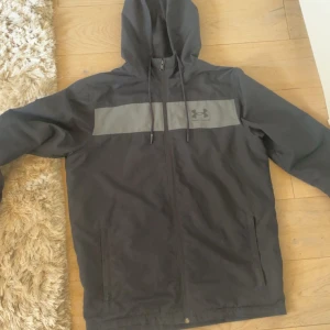 Svart vindjacka från Under Armour - Svart vindjacka från Under Armour med grått band över bröstet och logga. Jackan har huva med snörning, dragkedja framtill och två sidofickor. Lätt meshfoder på insidan och loose fit för en avslappnad stil.