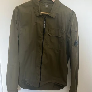 Olivgrön overshirt från C.P. Company - Säljer en olivgrön overshirt från C.P. Company med klassisk krage, bröstficka och ikonisk linsdetalj på ärmen. Jackan har dragkedja framtill och är tillverkad i ett lätt syntetmaterial.