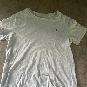 Grå t-shirt från Polo Ralph Lauren storlek 130cm - Snygg grå t-shirt från Polo Ralph Lauren med klassisk rund hals och korta ärmar. T-shirten har en liten mörkblå Polo-logga broderad på bröstet. Tillverkad i mjuk bomull som är skön mot huden och passar perfekt till jeans eller shorts. 