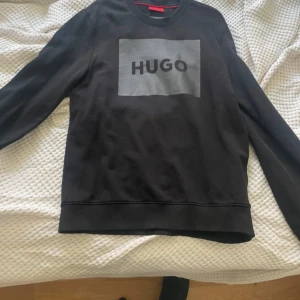 Svart sweatshirt från Hugo Boss - Svart sweatshirt från Hugo Boss med stor grå logga på bröstet. Tröjan har rund halsringning, långa ärmar och ribbade muddar. Perfekt för dig som gillar stilren och enkel design med en tydlig logga framtill.