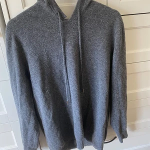 Kashmir tröja - Mysig grå hoodie i stickat material med huva och dragsnören. Tröjan har lång ärm och ribbade muddar vid ärmslut och nederkant. Perfekt för kyliga dagar när du vill vara både bekväm och snygg.