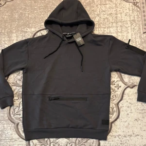 Mörkgrå hoodie från Much More - Snygg mörkgrå hoodie från Much More med huva och svarta dragsnören. Tröjan har en praktisk ficka med dragkedja framtill och en extra ficka med dragkedja på ena ärmen. Perfekt för dig som gillar stilren och funktionell streetwear. Helt ny, köpte för 40 euros. 