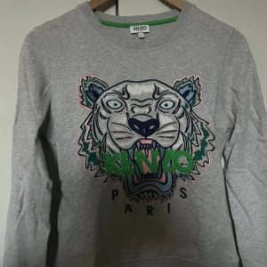 Grå Kenzo sweatshirt med tigertryck - Säljer en grå sweatshirt från Kenzo med ikoniskt broderat tigerhuvud i flera färger och grönt Kenzo-tryck på bröstet. Tröjan har rund hals, långa ärmar och ribbade muddar. Perfekt statement-piece för dig som gillar streetstyle.