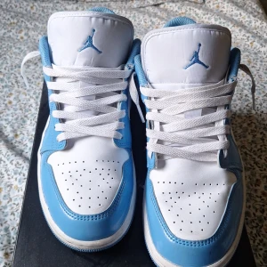 Nike Air Jordan 1 Low UNC blå/vit - Nike Air Jordan 1 Low i klassiska UNC-färger. Skorna har vit bas i läder med blanka ljusblå overlays och matchande swoosh. Broderad Air Jordan-logga på hälen och blå Jumpman på plösen. Lågt skaft, vita snören och perforerad tå för extra vibe. Pris kan gå ner vid snabb äffär