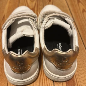 Vita sneakers från Björn Borg med guld - Snygga vita sneakers från Björn Borg med gulddetalj på hälen och praktiska kardborreband. Skorna har rund tå, platt sula och är tillverkade i syntetmaterial. Perfekta för dig som gillar stilrena och sportiga skor med en touch av guld.