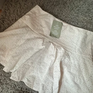 Vit broderad kortkjol från H&M - Superfin vit kortkjol från H&M med broderade blommor och lätt utsvängd passform. Kjolen är gjord av 20% återvunnen bomull och 64% återvunnen polyester, vilket gör den både snygg och hållbar. Perfekt för en fräsch och somrig look!