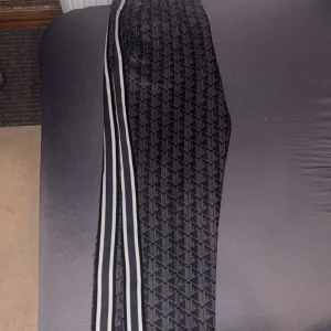 Svarta mönstrade byxor från Lacoste - Säljer ett par svarta byxor från Lacoste med grått geometriskt mönster och vita ränder längs sidorna. Byxorna har resår i midjan och en sportig vibe, perfekta för chill eller streetwear. Klassisk Lacoste-logga på höften.