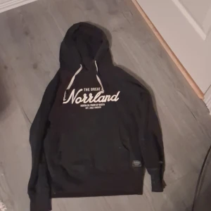 Svart hoodie från SQRTN Company - Svart hoodie från SQRTN Company med vit text 'The Great Norrland' på bröstet. Tröjan har huva med vita snören och klassisk passform. Perfekt för dig som gillar streetwear och vill ha en stilren look med norrländsk vibe.