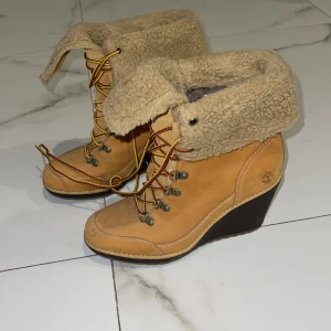 Timberland kängor med klack - Beiga Timberland skor med klack och snörning. Utsida i skinn och insida fodrad med mjuk teddy. Klassiska gula och röda snören samt vikbar krage. Perfekta för höst och vinter. Storlek 37,5