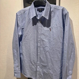 Blå skjorta från Ralph Lauren - Klassisk blå skjorta från Ralph Lauren med broderad logga på bröstet. Skjortan har lång ärm, knappar framtill och en stilren krage. Perfekt för dig som gillar en tidlös och clean look.Skulle säga att den passar också M