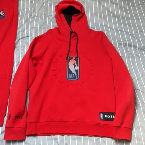 NBA x BOSS hoodie - Snygg röd hoodie från BOSS i samarbete med NBA. Stor NBA-logga framtill med BOSS-embossing runtom, svarta snören och patch nertill. Hoodien har lång ärm, huva med svart insida och är gjord i mjuk bomull. Perfekt för dig som gillar basket och streetwear.