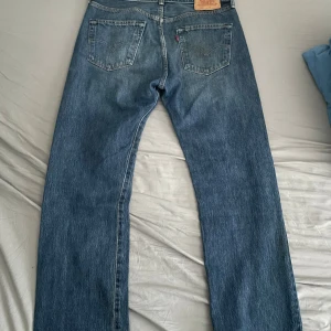 Levi's 501 blå jeans W33 L30 - Klassiska Levi's 501 jeans i blå tvätt med raka ben och normal passform. Jeansen har fem fickor, knappgylf och den ikoniska läderpatchen bak i midjan. Perfekta för dig som gillar tidlös stil och vill ha ett par schyssta jeans i bomull.