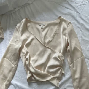 Champagnefärgad omlottopp från H&M - Snygg champagnefärgad omlottopp från H&M i satinliknande material. Toppen har lång ärm, v-ringning och en smickrande omlottdesign som ger en elegant look. Använd en gång och säljs pga den inte kommer till användning🥰