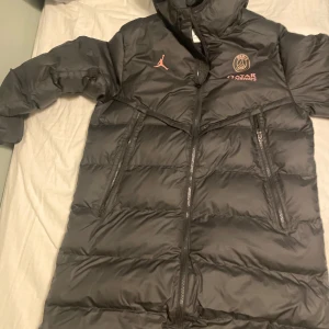 Svart PSG x Jordan dunjacka M - ( Kvitto tillkommer )Svart lång dunjacka från Jordan och PSG med rosa loggor och Qatar Airways-tryck. Jackan har huva, två stora dragkedjefickor och är vadderad för extra värme. Perfekt för kalla dagar och snygg streetstyle.