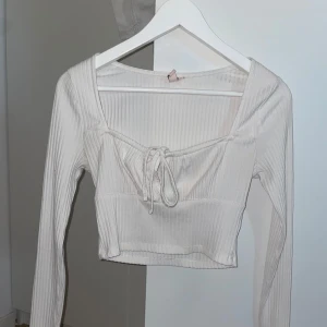 Vit ribbad croppad topp från Nelly - Säljer min vita croppade topp från Nelly med långa ärmar och fyrkantig ringning. Toppen är ribbad och har en söt knytning framtill som ger en extra detalj. Den är i storlek S och i mycket bra skick.