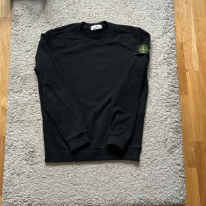 Svart sweatshirt från Stone Island - Svart långärmad sweatshirt från Stone Island med klassisk rund hals och ikonisk patch på vänster ärm. Tröjan är i mjuk bomull och har ribbade muddar vid ärmslut och nederkant. Perfekt för dig som gillar clean streetwear med premiumkänsla. Skicka gärna prisförslag! Bara på testad. Vet inte om den är äkta.