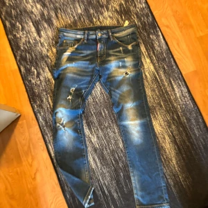 Blå  jeans från Dsquared2 - Snygga blå jeans från Dsquared2 med slitna detaljer och coola hål på benen. Modellen är klassisk femficksdesign och tydliga kontrastsömmar. Jeansen har en brun läderpatch med Dsquared2-logga bak och en röd logotag vid gylfen. Perfekt för dig som gillar streetstyle.