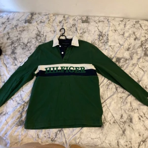 Grön pikétröja Tommy Hilfiger - Säljer en grön långärmad pikétröja från Tommy Hilfiger i klassisk fit. Tröjan har vit krage, mörkblå och vit rand över bröstet med stor Hilfiger-logga, samt en vit detalj på ena ärmen. Perfekt för dig som gillar sportig och preppy stil.