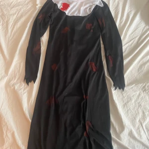 Halloween klänning med blod - En svart klänning med vita detaljer vid halsen och röda blodfläckar över hela klänningen. Långa ärmar med trasiga kanter ger en dramatisk look, perfekt för halloween eller temafest. Materialet känns lätt och stretchigt. Finns ingen lapp kvar där storleken står men skulle tro xs/s.