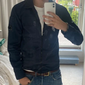 Mörkblå overshirt från C.P. Company - Säljer en mörkblå overshirt från C.P. Company med klassisk krage, dragkedja framtill och en ficka på bröstet. Ärmen har den ikoniska linsen på sidan. Perfekt för lager-på-lager och snygg till jeans.