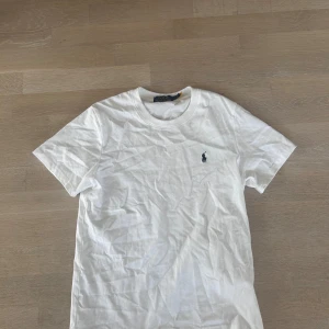 Vit t-shirt från Polo Ralph Lauren - Vit t-shirt från Polo Ralph Lauren med klassisk rund hals och korta ärmar. T-shirten har den ikoniska broderade Polo-loggan i mörkblått på bröstet. Tillverkad i mjuk bomull för skön passform och clean look.