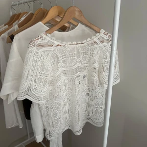 Vit spetsblus med kort ärm - Superfin vit blus i spets med korta ärmar och rund hals. Blusen har ett detaljerat broderat mönster och är lätt transparent, perfekt för att skapa en romantisk och trendig look. Passar snyggt till både jeans och kjol.