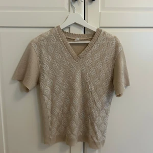 Beige stickad topp med v-ringning - Snygg beige stickad topp med korta ärmar och v-ringning. Framsidan har ett diskret mönster i vitt som ger en retrovibe. Perfekt till jeans eller kjol för en chill och stilren look.