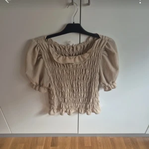 Beige blus med puffärm och smock - Supersöt beige blus med smockad kropp och puffiga korta ärmar. Blusen har en bred, rundad halsringning och volangdetaljer vid ärmslut och nederkant. Perfekt för dig som gillar romantisk och trendig stil.
