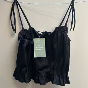 Svart ribbad linnetopp från H&M - Supersöt svart ribbad linnetopp från H&M med smala axelband som knyts upptill och volangkant nertill. Tillverkad av 58% återvunnen polyester. Perfekt för en chill och trendig sommarlook.