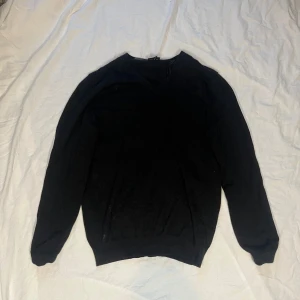 Svart stickad tröja Galvin Green - Svart stickad tröja från Galvin Green med v-ringad hals och långa ärmar. Enkel och stilren design som passar perfekt till jeans eller chinos. Tröjan har ribbade muddar vid ärmslut och nederkant för en snygg passform.