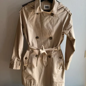 Beige trenchcoat med huva från Zara Kids - Stilren beige trenchcoat från Zara Kids i storlek 164 (13-14 år) med huva, dubbelknäppning och bälte i midjan. Jackan har långa ärmar, snygga knappar och praktiska fickor framtill. Perfekt för vår och höst. Sparsamt använd och i jättebra skick🤩✨