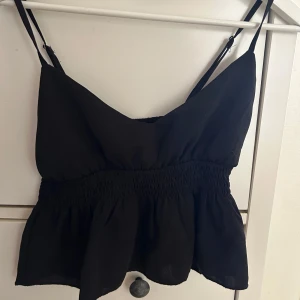 Svart volangig linnetopp - Supersöt svart linnetopp med smala axelband och volangig nederdel. Toppen har smockad midja som ger en snygg passform och en luftig känsla. Perfekt att styla med jeans eller kjol för en trendig look.