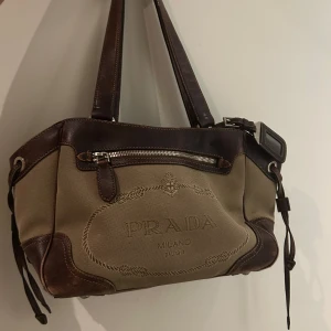 Prada handväska  - Säljer en snygg vintage handväska från Prada i beige canvas med mörkbruna skinn-detaljer. Väskan har dubbla handtag, dragkedja fram och bak samt broderad Prada-logga på framsidan. Insidan är fodrad med mönstrat tyg och har klassisk Prada-label. Den är väl använd, därav lite slitage på skinnet, men inget som ”påverkar” väskan. Äktahetsbevis finns (se bilder) men inte dustbag eller kvitto i pappersformat. 