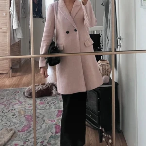 Ljusrosa kappa från Mango - Säljer en stilren ljusrosa kappa från Mango i storlek S. Kappan har dubbelknäppning med svarta knappar, klassisk krage och två fickor framtill. Materialet är en mjuk ullblandning som ger en lyxig känsla. Perfekt för dig som gillar clean och chic look.