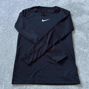 Svart långärmad Nike Dri-Fit tröja - Svart långärmad tröja från Nike med Dri-Fit-teknologi. Tröjan har en stilren look med Nike-logga på bröstet och är tillverkad i ett lätt och snabbtorkande syntetmaterial, perfekt för träning eller sportiga dagar.