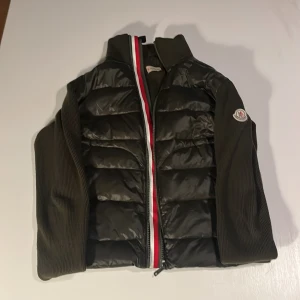 Svart pufferjacka från Moncler - Säljer en svart pufferjacka från Moncler med stickade ärmar och sidor. Jackan har en snygg dragkedja med röd, vit och blå rand samt Moncler-logga på ärmen. Perfekt för dig som vill ha en stilren och sportig look. Har också triangeln hemma. Kom pm priset kan diskuteras 👌