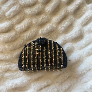 Svart axelväska med paljetter och guld - Unik svart axelväska i halvmåneform med glittriga paljetter och vertikala ränder i guld och brons. Väskan har en smal axelrem och stängs med magnetlås. Perfekt för att lyfta din outfit med extra bling och cool vintagekänsla.