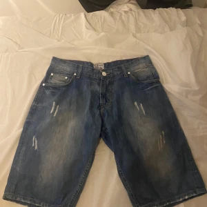Blå jeansshorts från Fashion Factory - Snygga blå jeansshorts från Fashion Factory i klassisk femficksmodell. De har slitna detaljer framtill och en lite längre passform som ger en avslappnad vibe. Perfekta för sommardagar när du vill ha en cool och enkel stil.