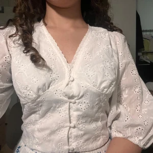 Vit broderad blus med knappar - Superfin vit blus med broderade blommor och v-ringning. Blusen har puffiga trekvartsärmar, klädda knappar framtill och spetskant vid halsen. Perfekt för dig som gillar romantisk och söt stil.