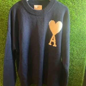 Mörkblå stickad tröja från Ami - Snygg mörkblå stickad tröja från Ami Alexandre Mattiussi med ett stort vitt hjärta och bokstaven A på bröstet. Tröjan har rund hals, långa ärmar och ribbade muddar. Perfekt för dig som gillar stilrena detaljer och vill sticka ut med en ikonisk design.