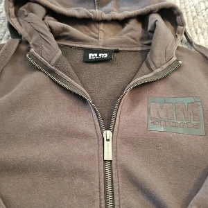 Brun hoodie från MM Sports med dragkedja - Mysig brun hoodie från MM Sports med huva och hel dragkedja framtill. Tröjan har ett stort tryck med MM Sports-logga på bröstet och praktiska fickor. Perfekt för chill eller träning, i mjukt material och med ribbade muddar.